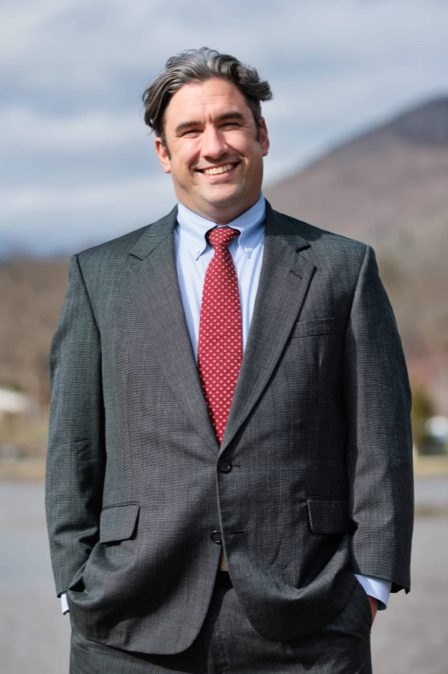 Benjamin, a lawyer at Stone & Christy, poses in a suit with a red and white polka dot tie for a picture with the mountains in the background.