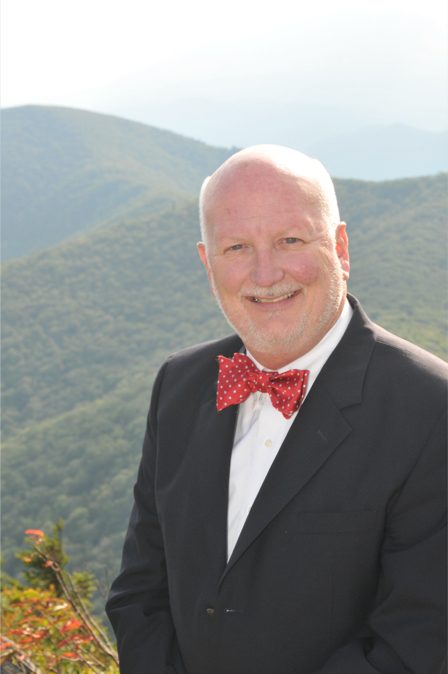 William, a lawyer at Stone & Christy, poses in a suit with a red bow tie with white polka dots for a picture with the mountains in the background.